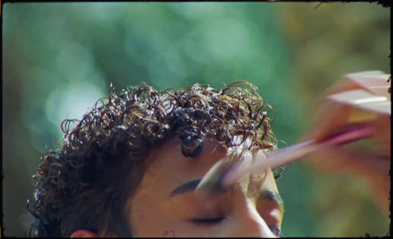 A persons hand using a brush to apply makeup to the face of a young man who has his eyes closed