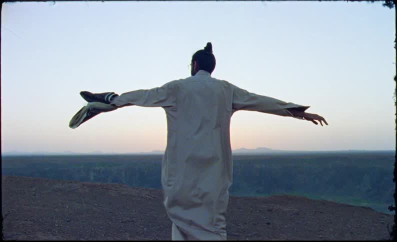 A man in a religious garb climbs a hill he stretches his arms out as he is buffetted by the wind as he reaches the top