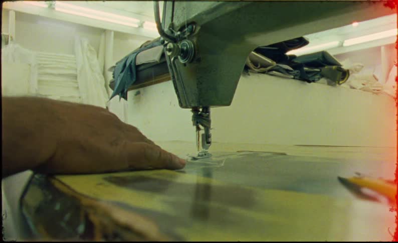 A mans hands guide fabric through a large industrial sewing machine in a dimly lit workshop