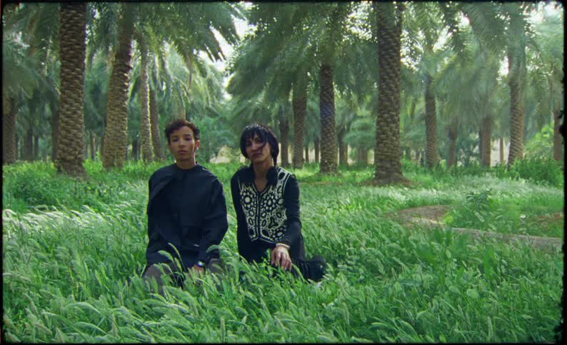 A young man and woman are sitting on the grass and looking straight ahead as the woman brushes her hair out of her face