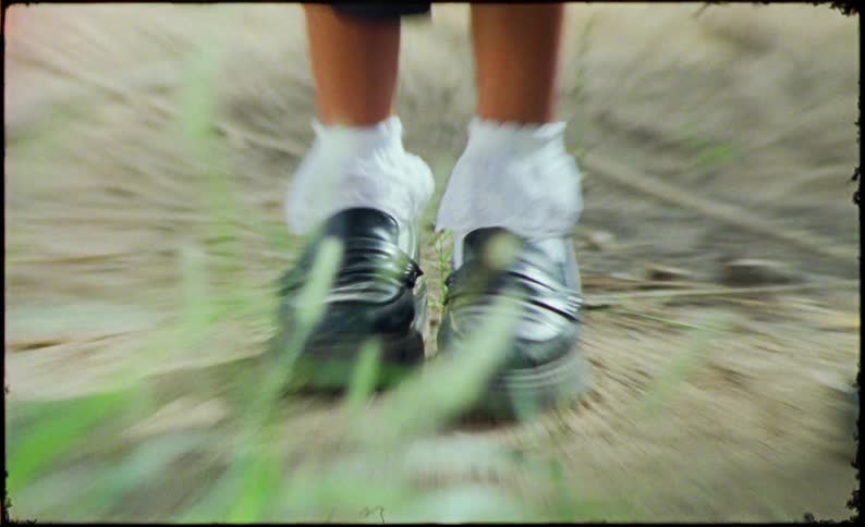 Two pairs of legs wearing black skirts white socks and black loafers standing on a dirt path