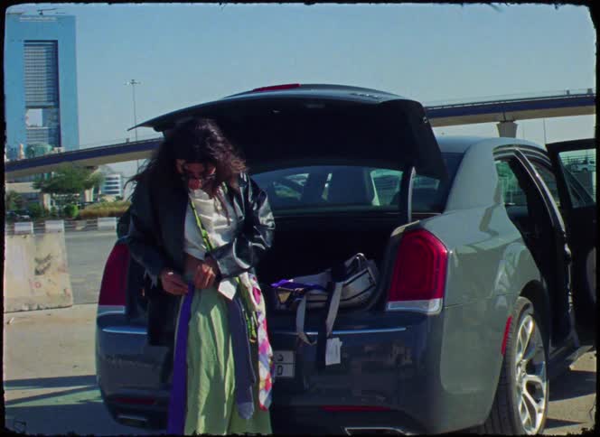 A woman standing near an open car trunk is adjusting her clothing wearing a green skirt a leather jacket and sunglasses with an urban backdrop featuring an overpass and modern buildings