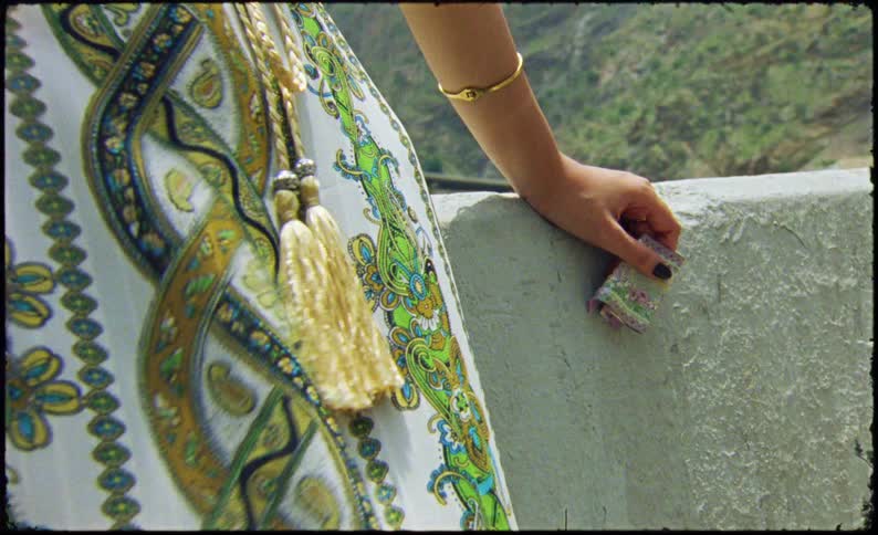 A womans hand adorned with a gold bangle holding a colorful item against a patterned fabric with intricate designs and tassels set against a backdrop of a mountainous landscape