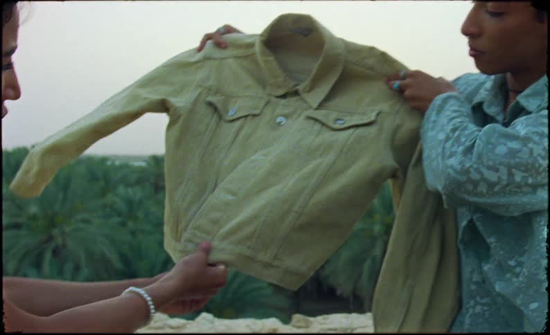 A person with short curly hair holds up a beige denim jacket wearing a patterned light blue shirt set against a soft outdoor background
