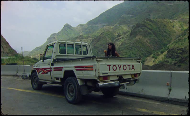 Woman eating a fruit next to a small truck stopped on the highway with a background surrounded by greenish mountains