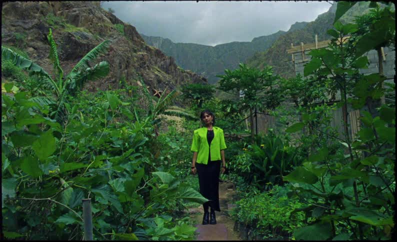 A woman wearing a bright green jacket and black dress stands on a narrow path surrounded by lush dense vegetation