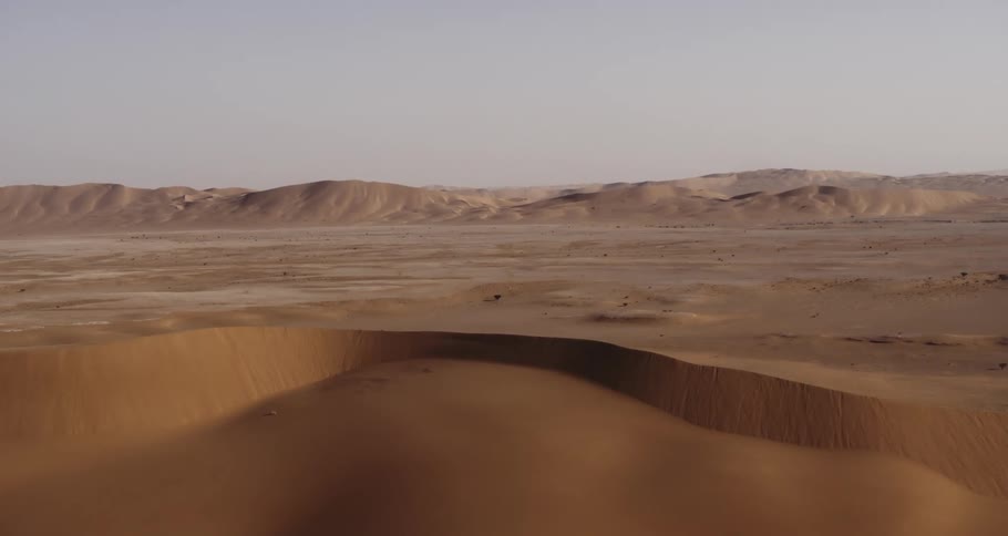 A drone moves over a dune, revealing a vast desert landscape featuring rolling sand dunes and an expansive horizon
