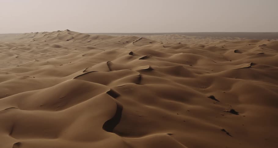 An expansive desert landscape with undulating sand dunes stretches into the horizon under a hazy sky
