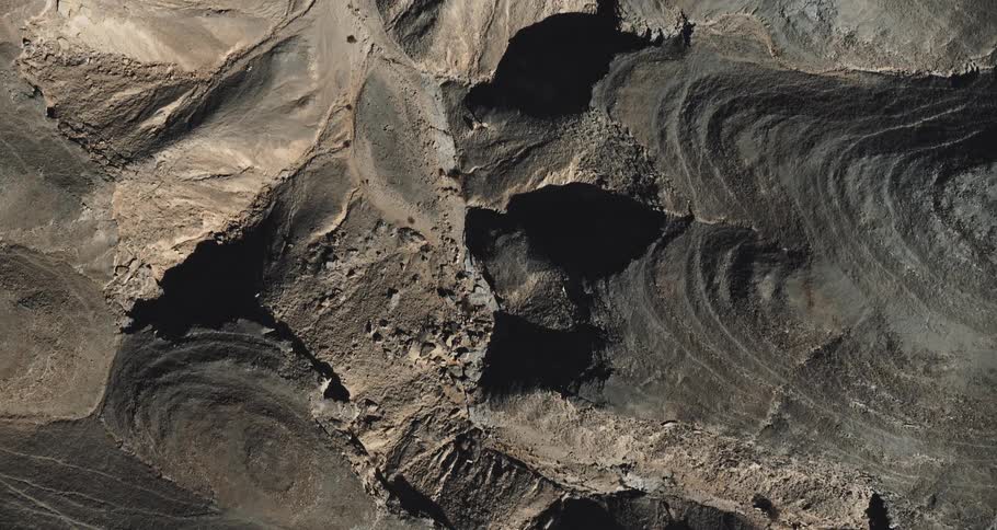 An aerial view of a dark rugged barren landscape with deep crevices and eroded rock formations