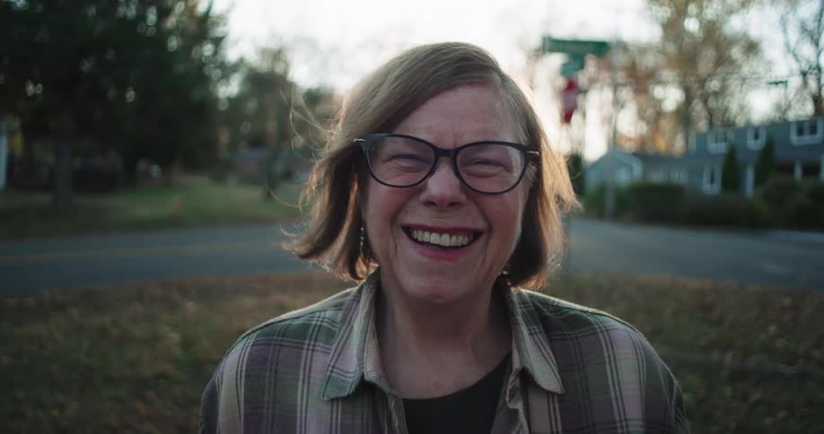 A cheerful woman with glasses and short hair smiling warmly in an outdoor suburban setting during what appears to be late afternoon with soft sunlight and trees in the background