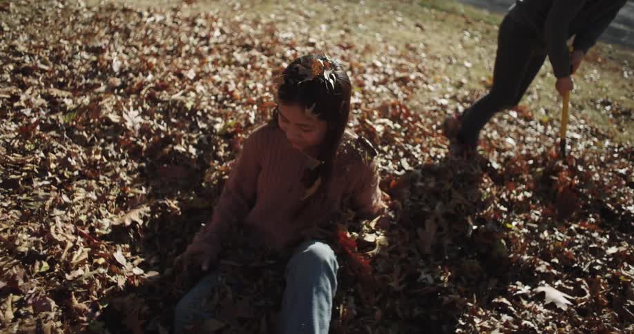 Girl happily sitting on a pile leaves tossing in the air as her brother rakes leaves on her