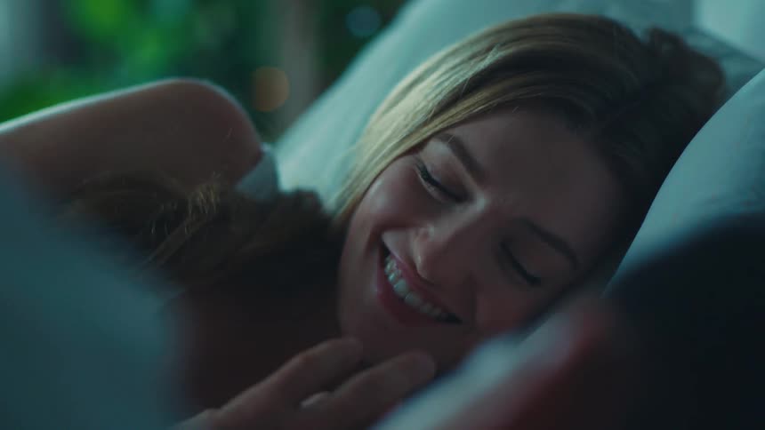 A blonde woman and a man lie in bed touching hands in the bedroom
