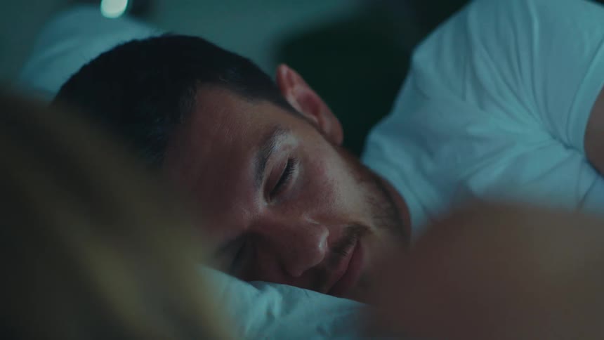 A man and a woman are lying in bed touching hands and engaged in conversation
