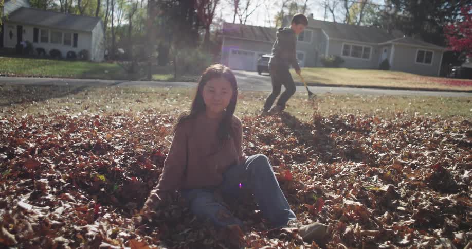A young girl sits joyfully in a pile of autumn leaves gesturing playfully while another person rakes leaves in the background near a suburban street lined with houses and trees