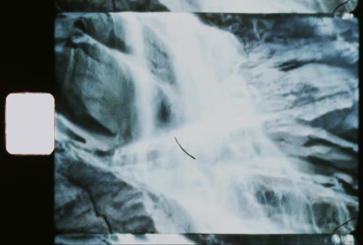 A person in an orange jacket stands near a powerful waterfall cascading down a rocky cliff face