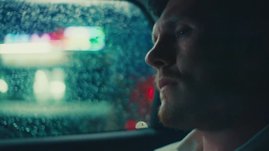 A pensive man wearing a white shirt travels in a car as it drizzles at night in the city