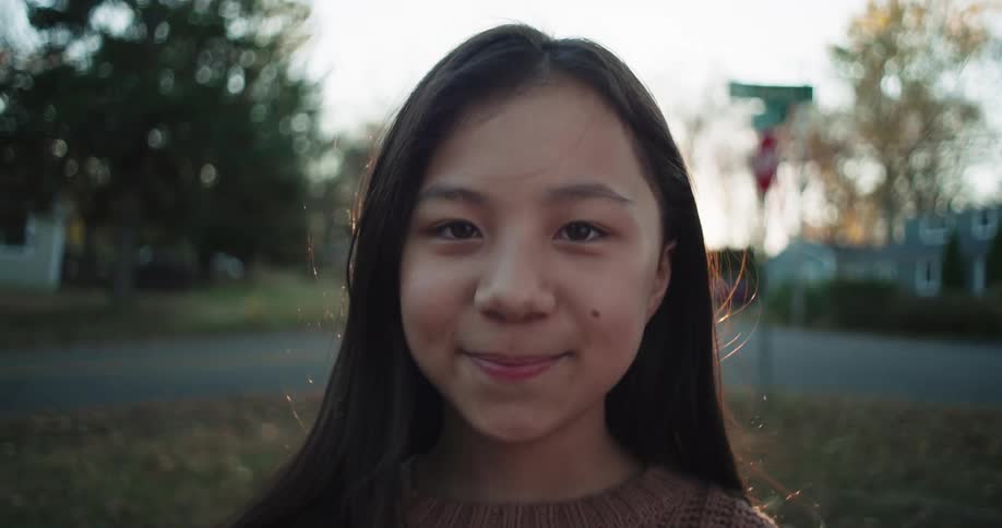 A girl with long hair stands outdoors in a residential area looking at the camera