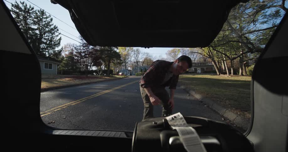 A man seen from the back of an open car trunk reaching in to retrieve or place a suitcase on a suburban street