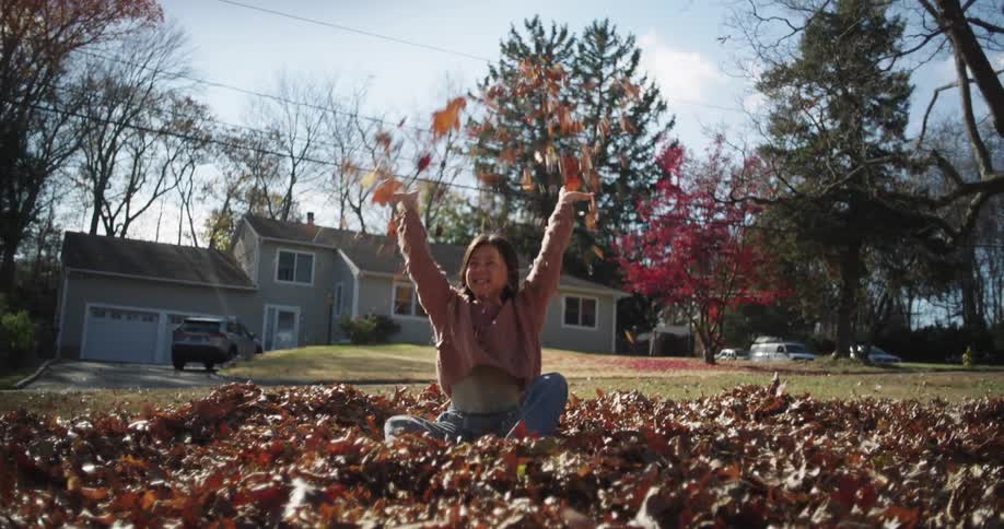 A girl is sitting in a pile of fallen leaves in a yard joyfully throwing leaves into the air