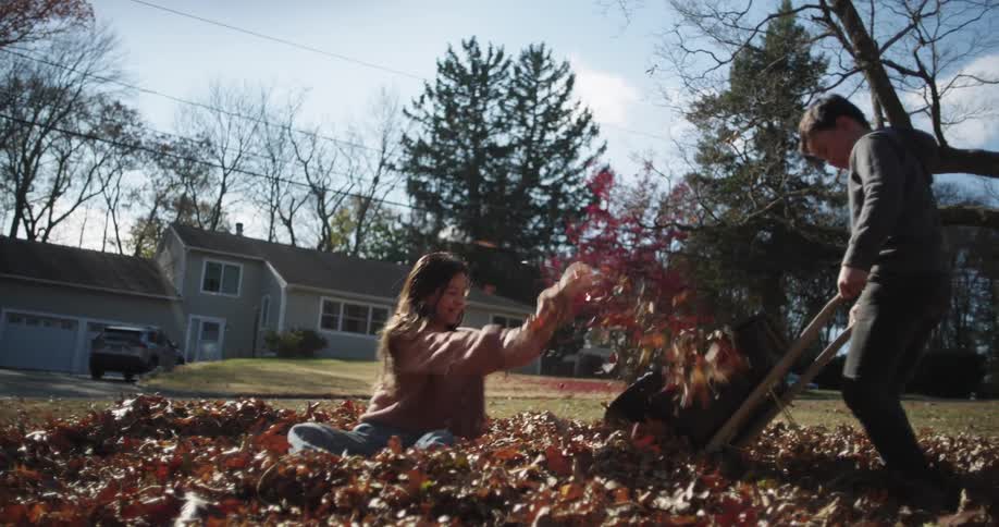 Siblings playing in a pile of leaves throwing leaves in a wheelbarrow