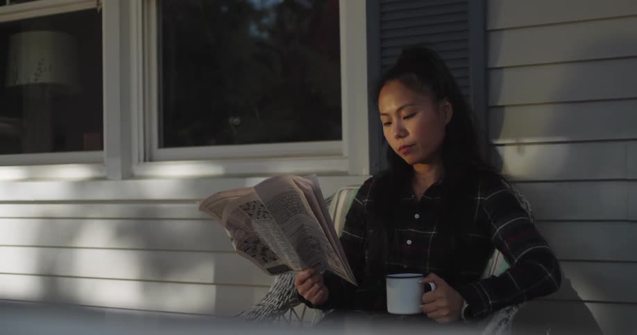 Woman sitting drinking coffee and reading a newspaper on the porch of her house