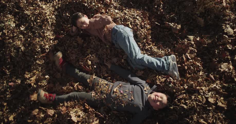 Two children are lying on a bed of autumn leaves gazing at each other with sunlight casting soft shadows across the scene and a natural playful atmosphere