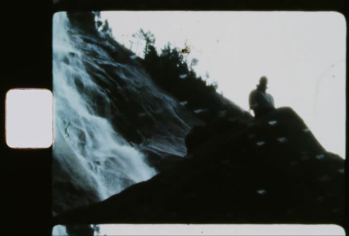 A person climbing a dark rock and there is a person already at the top looking and behind there is a waterfall with some water falling