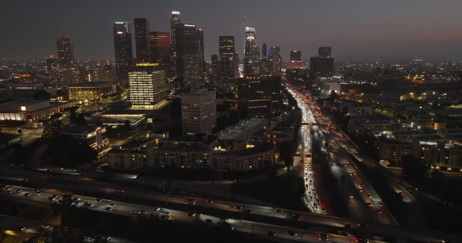 A nighttime aerial view of a bustling cityscape with illuminated skyscrapers and heavy traffic on a multilane highway
