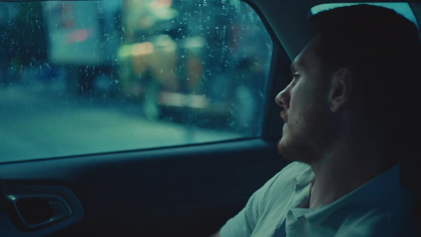 A man sits in a car at night looking pensively out the window with city lights twinkling in the background