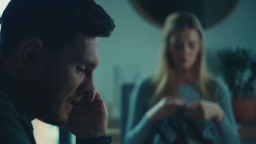 A man is speaking intently while a woman initially knitting a sweater suddenly looks up at him with wide surprised eyes