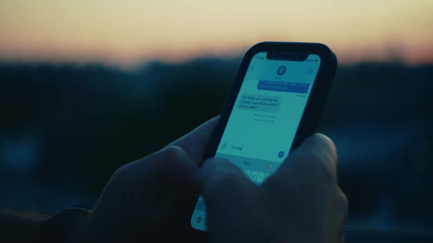 A mans hands typing on a smartphone against a blurred background of a sunset sky