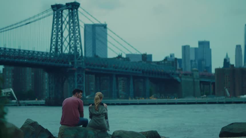 A couple sits by the waterfront gazing at a city skyline with a prominent bridge under a cloudy sky