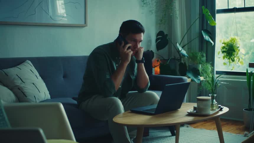 A man sits on his couch while talking on a smartphone and scrolling on his laptop