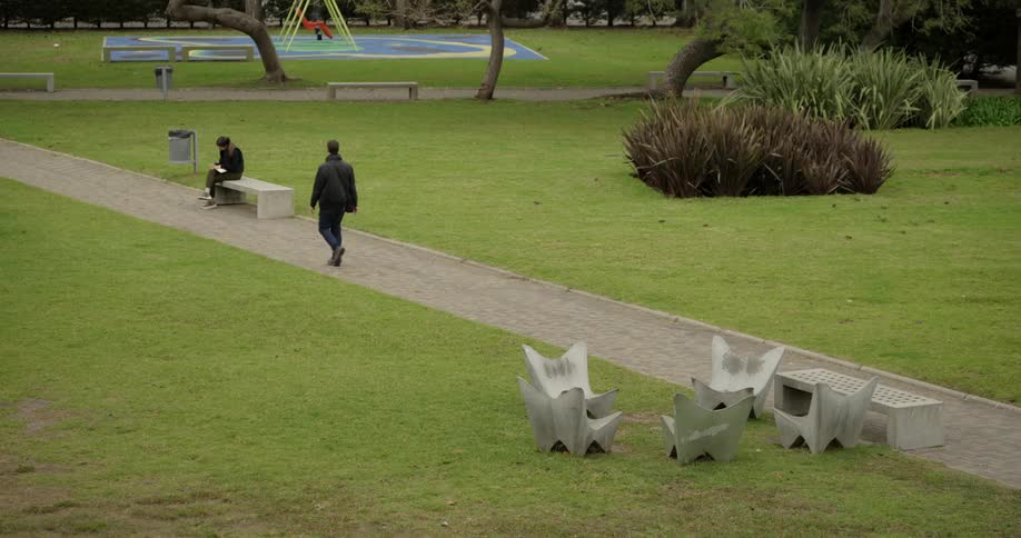Man walking through a public square and a woman in the background reading a book sitting on a bench