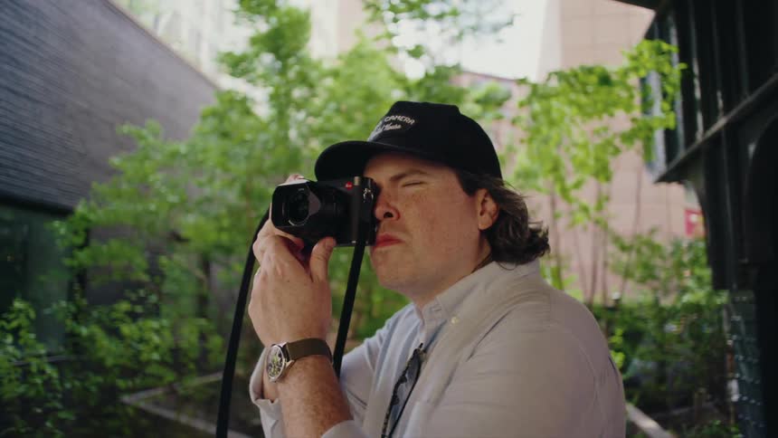 A man with a cap focusing through a camera outdoors surrounded by greenery