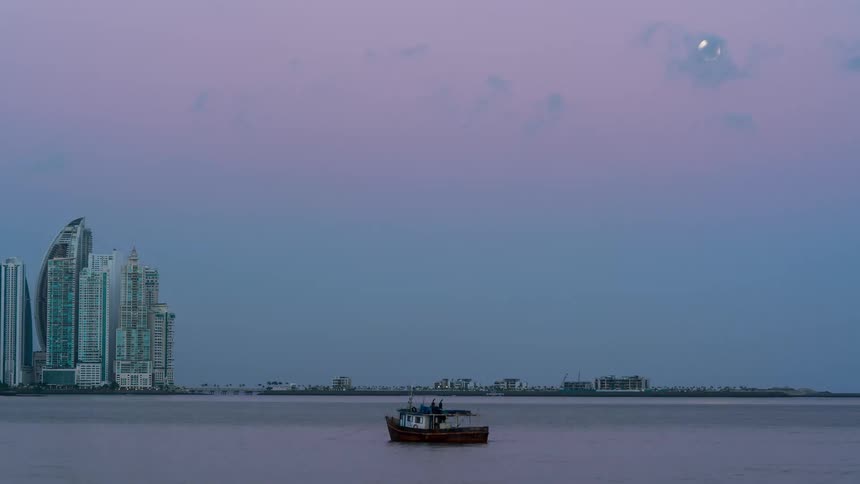 A small fishing boat floats in calm water with a modern cityscape and large moon in the background
