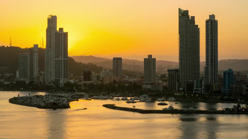 Boats maneuver into the marina of a city as the sun sets behind nearby mountains