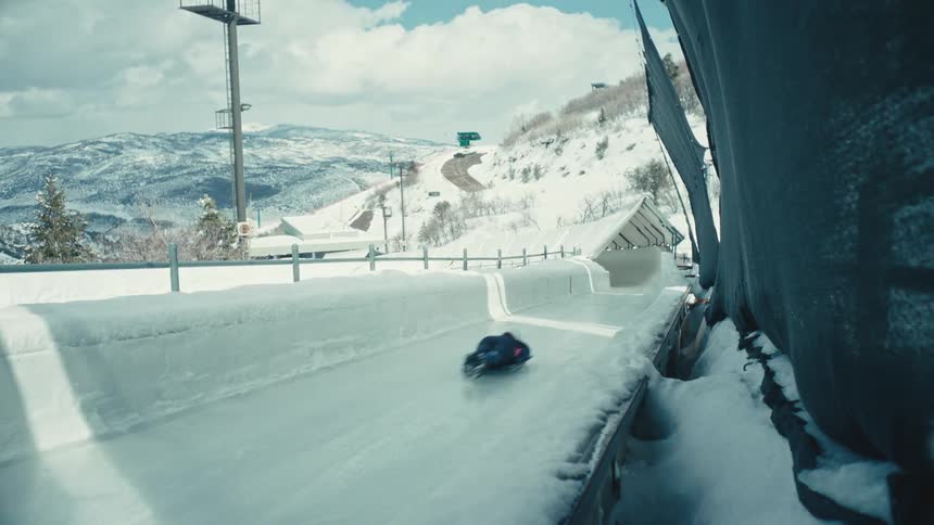 A skeleton racer speeds down an icy track viewed from a partially obscured perspective near the tracks edge with a snowy mountain landscape in the background