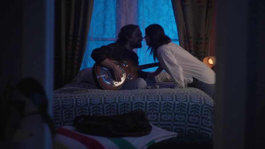 Man sitting on bed playing guitar and a woman giving him a kiss
