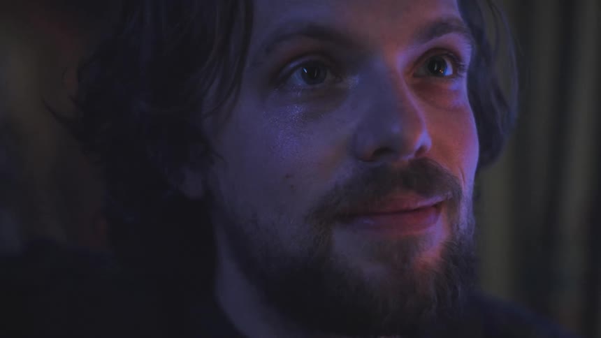 The face of a young bearded man with a small smile on his face and he is staring straight ahead while something is lighting up his face