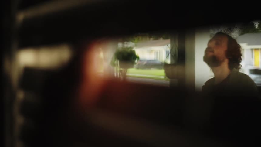 The face of a bearded man seen through a window blind opened by a person who is peeking inside