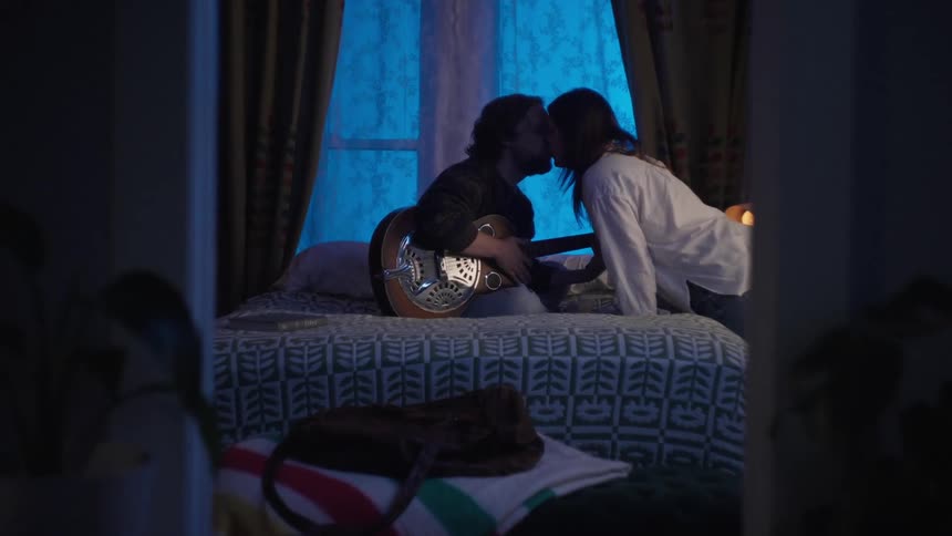 A man holding a guitar is smiling and sitting on a bed and then a woman appears and kisses him