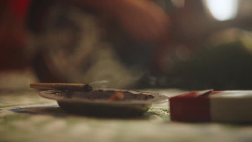 A persons hand placing a cigarette with smoke coming out of its tip on top of an ashtray