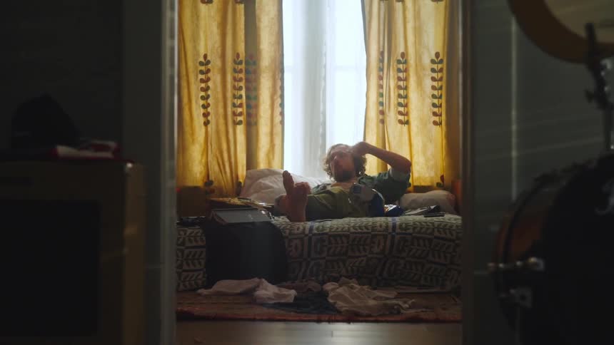 Sprawled across a rumpled bed in a room littered with clothes and takeout containers a man with long hair enjoys a bag of chips