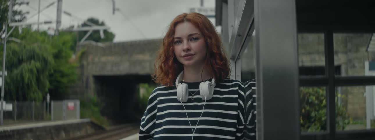 A woman with white headphones around her neck is standing outside against a metal structure