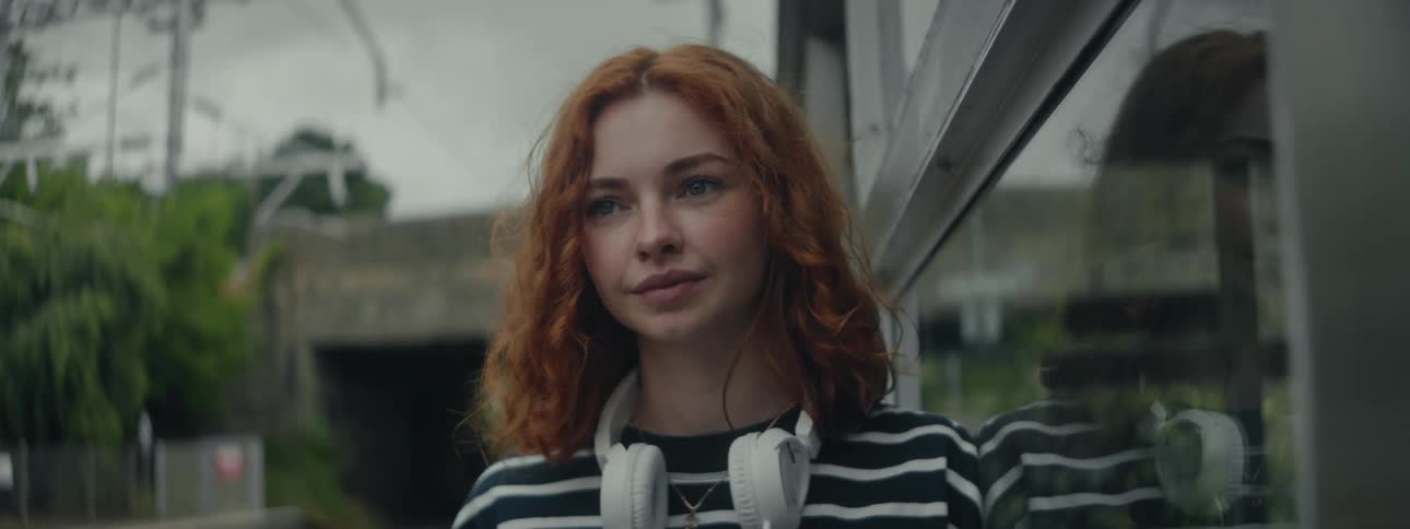 A woman with striking red hair and headphones stands near the train station