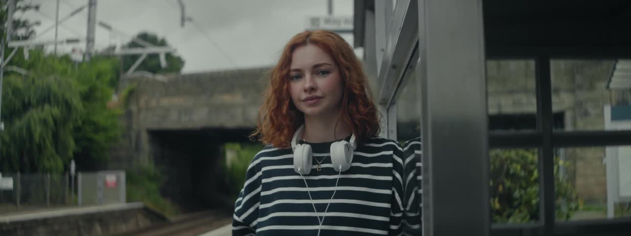 A woman with striking red hair black and white striped sweatshirt and headphones stands near the train station check the grammar