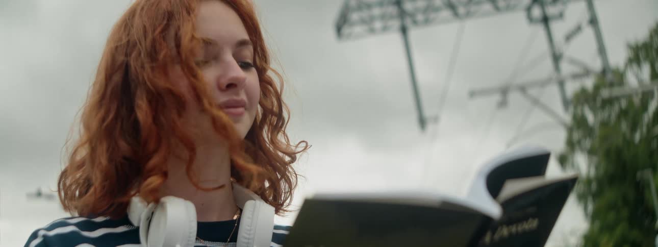 A young woman with red hair with headphones around her neck and she is reading a book that is in her hands