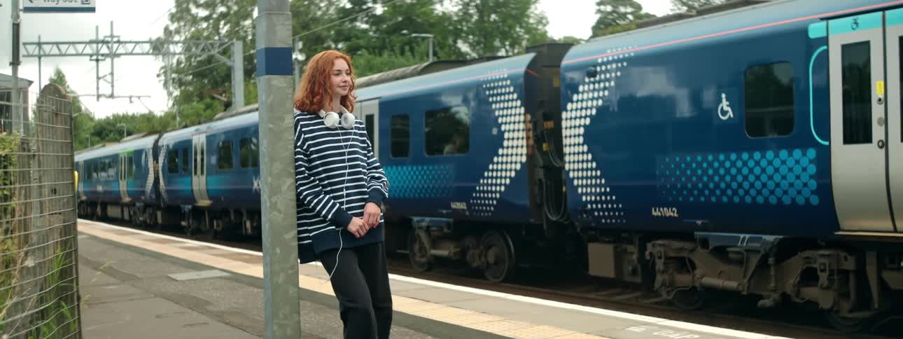 A young woman is outside leaning on a metal pole next to a train