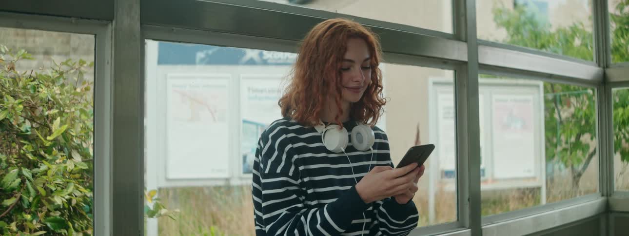 A young woman with curly red hair wearing a striped shirt and headphones around her neck smiles while looking at her phone